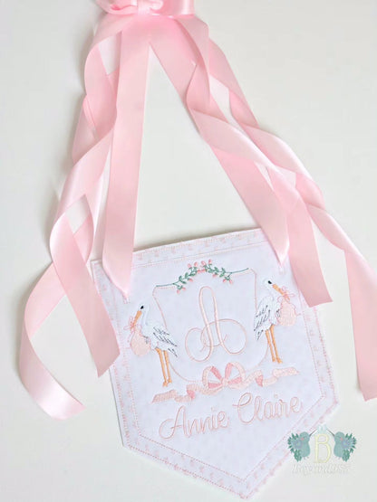Personalized baby girl door hanger with embroidered monogram, stork design, pink ribbon accents, and custom name embroidery.
