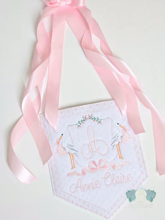 Personalized baby girl door hanger with embroidered monogram, stork design, pink ribbon accents, and custom name embroidery.
