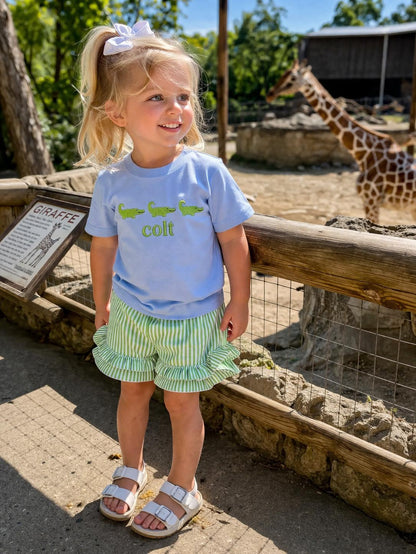 Young girl in a blue shirt and green shorts standing in front of a giraffe at a zoo.